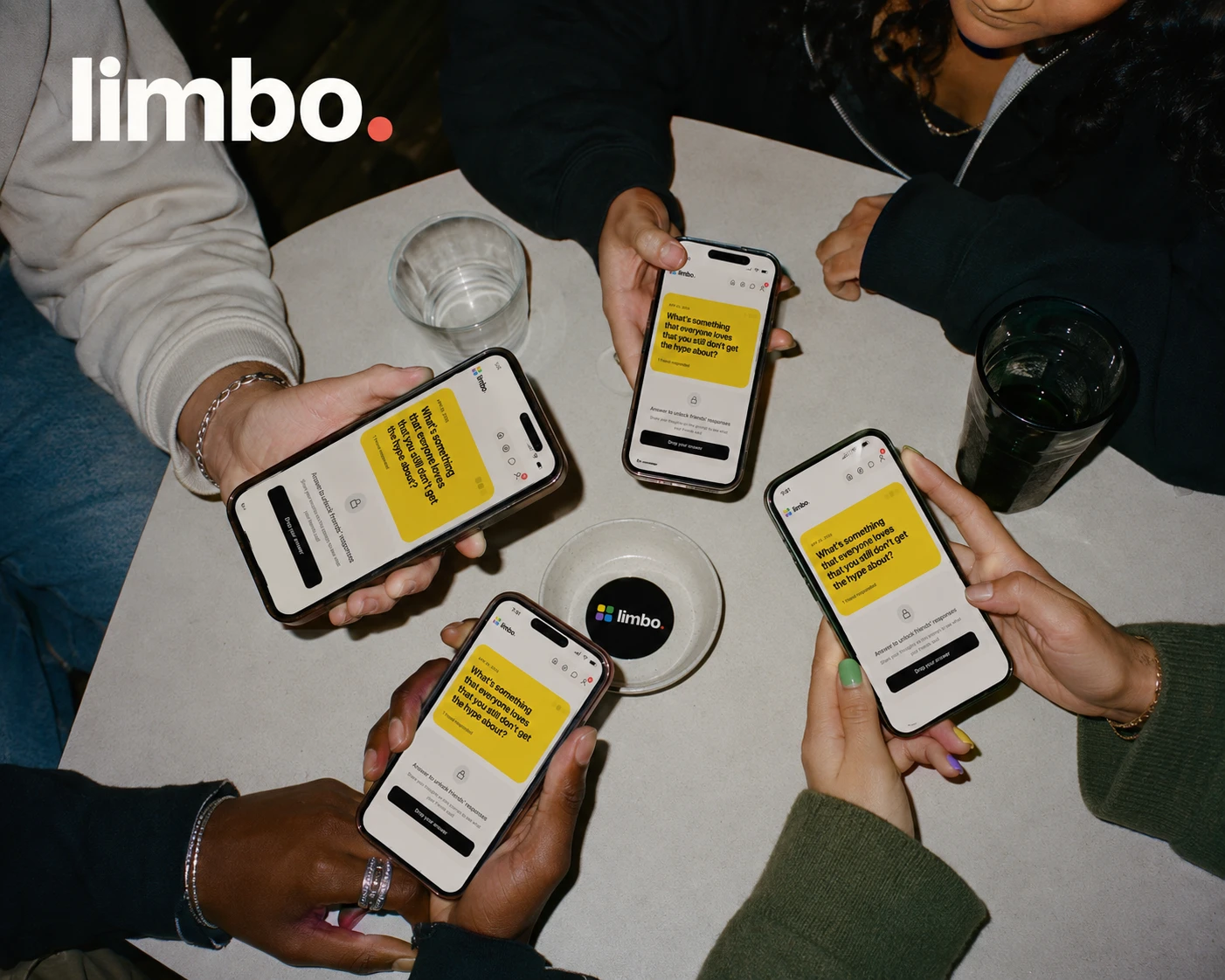 Four friends around a table, each holding a phone showing the same daily Limbo prompt — 'What's something that everyone loves that you still don't get the hype about?' — with a locked answer field.